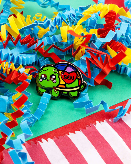 The Speshell Educational Needs enamel pin by Innabox shown on shredded colourful paper. The pin features a kawaii green tortoise with a rainbow shell and a red heart that says “SEN”, designed to promote awareness of special educational needs. Sold by Rebel Romance, a UK small business known for alternative gifts and fashion.