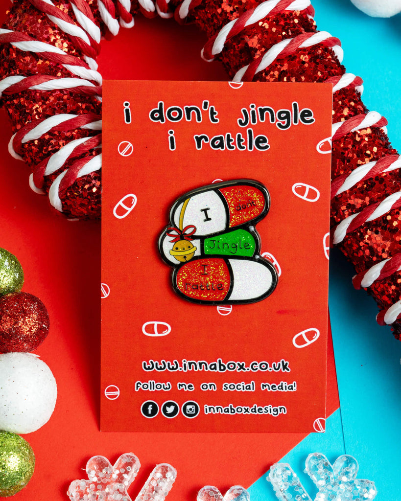 Festive enamel pin badge reading “I Don’t Jingle, I Rattle” designed by Innabox, featuring three glittery capsules with a jingle bell. Displayed on a red backing card with pills printed on it, surrounded by Christmas decorations. Sold by Rebel Romance, a UK-based alternative fashion and lifestyle brand.