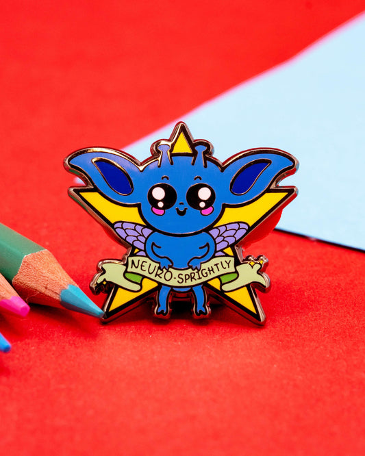 A close-up of a blue fantasy creature enamel pin with a yellow star and purple wings, displayed on a pink and orange backing card that reads “Neuro-Sprightly.” Designed by Innabox and sold by Rebel Romance, a UK-based alternative fashion and lifestyle brand