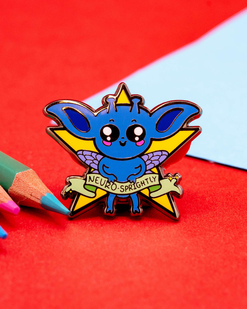 A close-up of a blue fantasy creature enamel pin with a yellow star and purple wings, displayed on a pink and orange backing card that reads “Neuro-Sprightly.” Designed by Innabox and sold by Rebel Romance, a UK-based alternative fashion and lifestyle brand