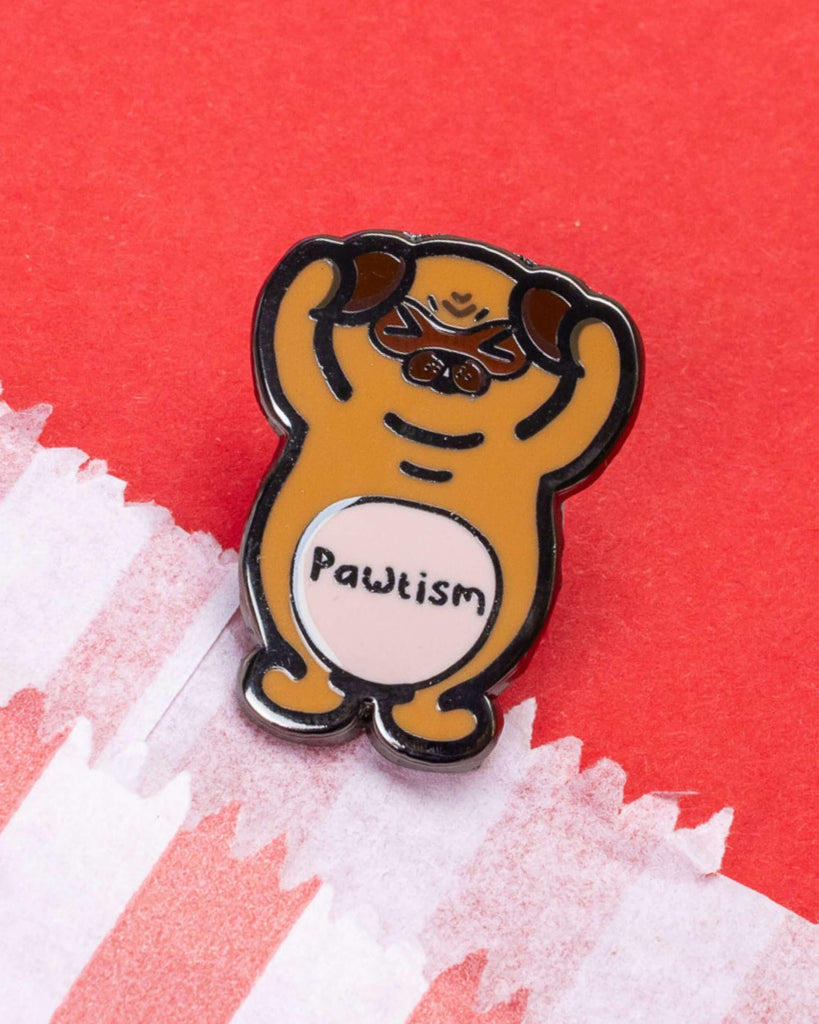A close-up of the Pawtism Dog Enamel Pin sold by Rebel Romance, a UK alternative lifestyle brand. The pin features a cartoon-style brown pug with its paws raised, a cute expression, and the word “Pawtism” on its tummy. Set against red paper and pastel stripe tissue, ideal for autism awareness and neurodiversity support.