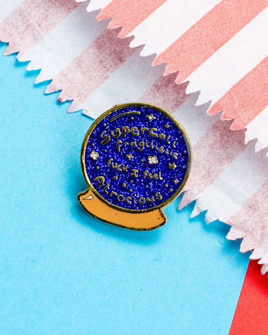 A glittery enamel pin shaped like a crystal ball, with the words "Supercalifragilistic fuck I feel Atrocious" written across a deep blue glitter background. The pin is placed on pastel blue paper with a striped pink and white paper partially visible.