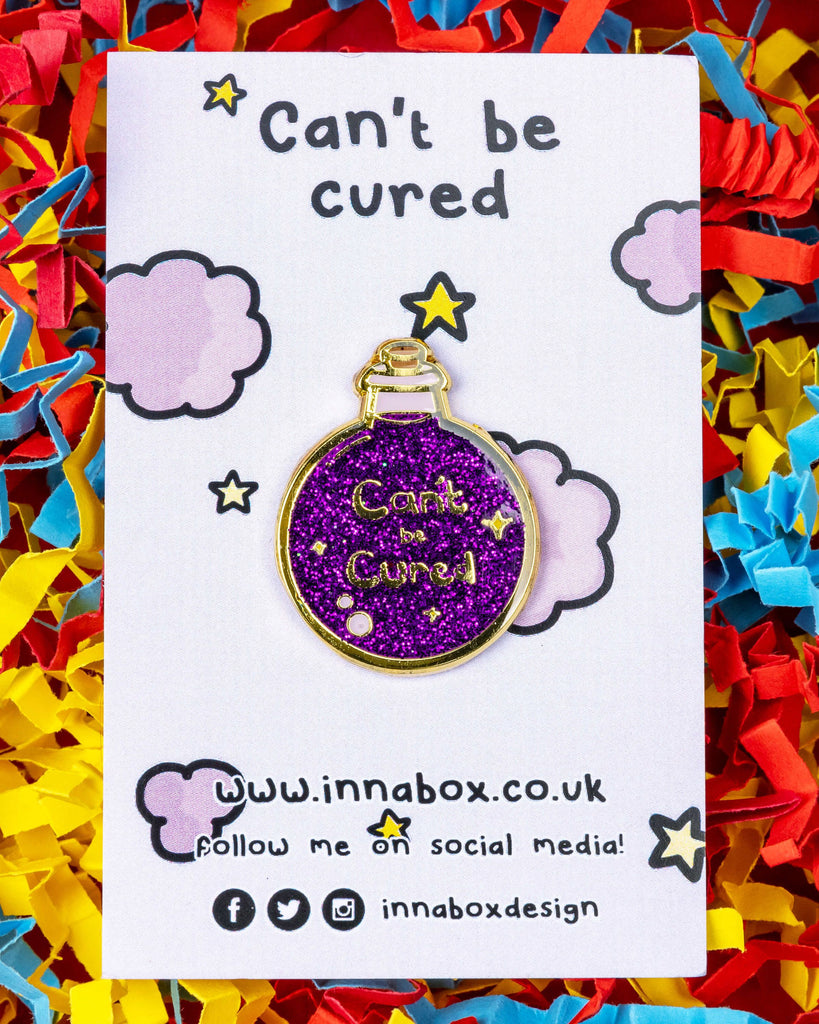 A glittery purple enamel pin by Innabox on a white card with pastel clouds and stars. The pin is shaped like a round potion bottle with gold trim and sparkles, and reads "Can't be Cured". Product from Rebel Romance, a UK small business celebrating alternative fashion and quirky accessories.