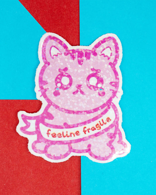 A glittery pink cat-shaped sticker from Rebel Romance with teary eyes and the phrase “feeline fragile” across its chest. The sticker is set against a bright blue and red background and designed by Innabox to raise awareness around chronic illness and hidden disabilities.