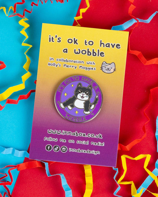 Enamel pin on rainbow gradient card featuring a black and white cat with the phrase "It’s OK to Have a Wobble" in purple and yellow. Designed by Innabox and sold by Rebel Romance, a small UK business celebrating alternative fashion, gifts, and self-expression.