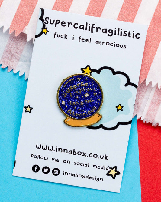 A glittery crystal ball enamel pin attached to a backing card that reads "Supercalifragilistic fuck I feel atrocious". The backing card is surrounded by soft pastel paper, and the pin sparkles with dark blue glitter and gold-toned edging.