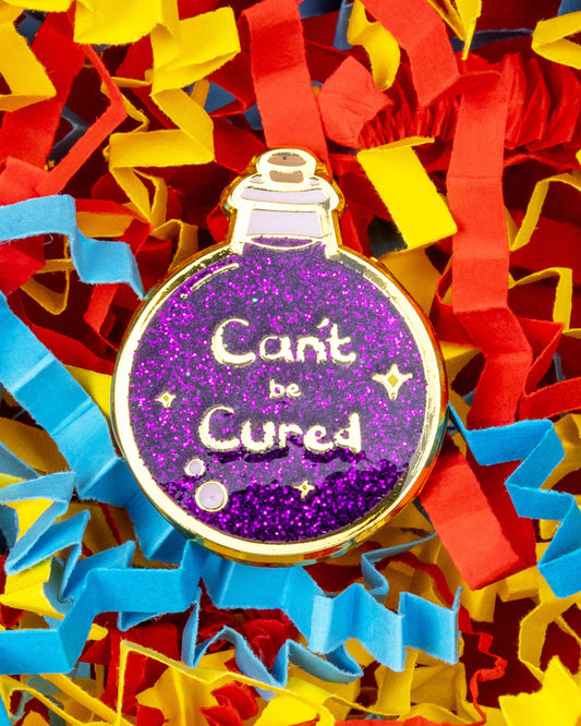 Close-up of a glittery purple potion bottle enamel pin reading "Can't be Cured", surrounded by colourful red, yellow and blue shredded paper. Designed by Innabox and sold by Rebel Romance, a UK-based alternative fashion and gift brand.