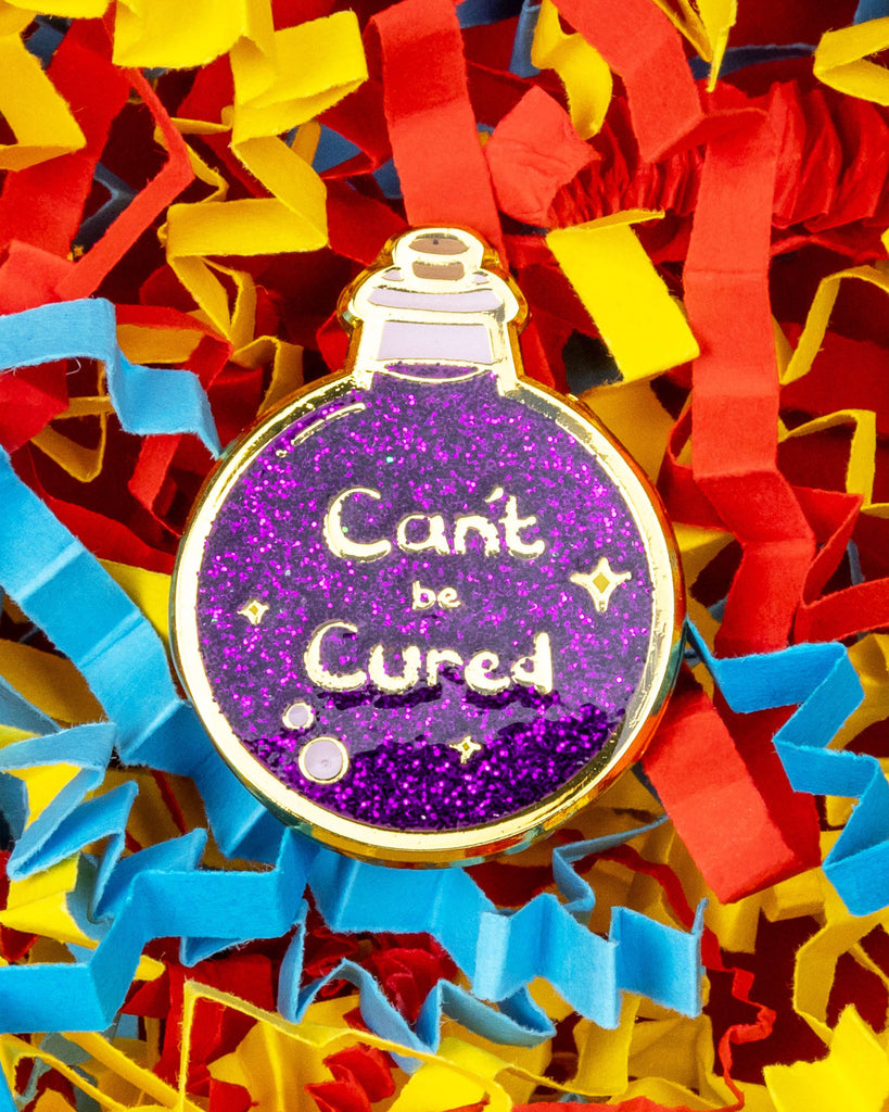Close-up of a glittery purple potion bottle enamel pin reading "Can't be Cured", surrounded by colourful red, yellow and blue shredded paper. Designed by Innabox and sold by Rebel Romance, a UK-based alternative fashion and gift brand.