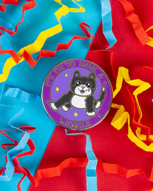 Close-up of the "It’s OK to Have a Wobble" cat enamel pin, designed by Innabox and available on Rebel Romance. The cute black and white cat sits on a purple circular pin with stars and text, surrounded by colourful confetti. Celebrates mental and physical health awareness.