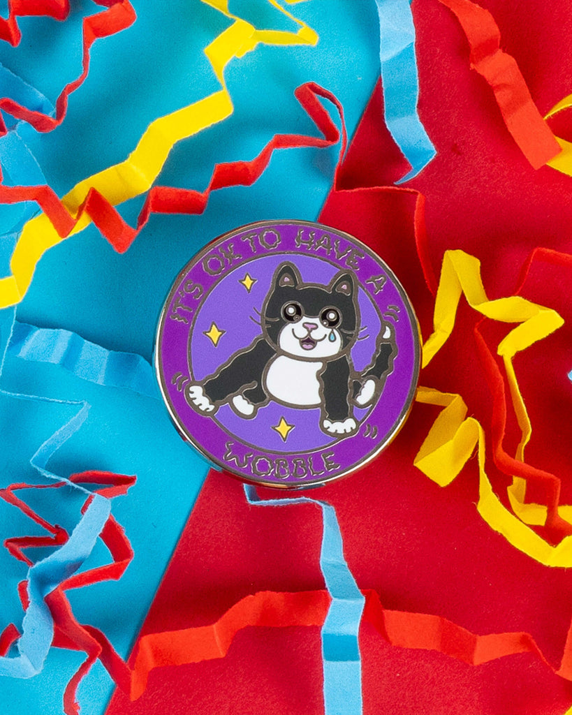 Close-up of the "It’s OK to Have a Wobble" cat enamel pin, designed by Innabox and available on Rebel Romance. The cute black and white cat sits on a purple circular pin with stars and text, surrounded by colourful confetti. Celebrates mental and physical health awareness.
