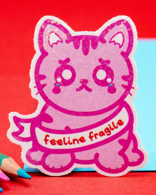 A glittery pink cat-shaped sticker from Rebel Romance with teary eyes and the phrase “feeline fragile” across its chest. The sticker is set against a bright blue and red background and designed by Innabox to raise awareness around chronic illness and hidden disabilities.