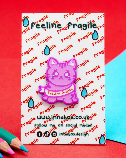 Photo of the Feeline Fragile enamel pin by Innabox on a backing card with a red and blue background. The pin features a pink cartoon cat with tears in its eyes and the phrase “feeline fragile” on a ribbon. Sold by Rebel Romance, a UK small business celebrating alternative fashion and neurodiverse pride.