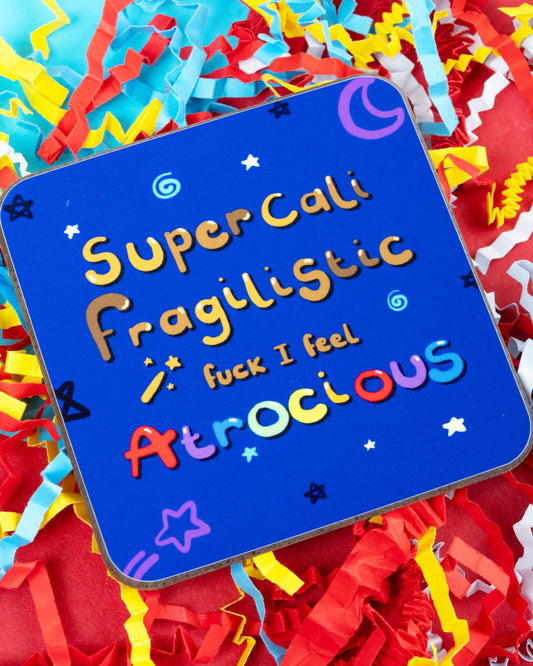 Close-up of the “I Feel Atrocious” coaster by Innabox on a bed of colourful shredded paper. The square wooden coaster features bright rainbow text on a dark blue night sky background with stars and moons. Sold by Rebel Romance, a UK indie brand for quirky, alternative gifts and homeware.