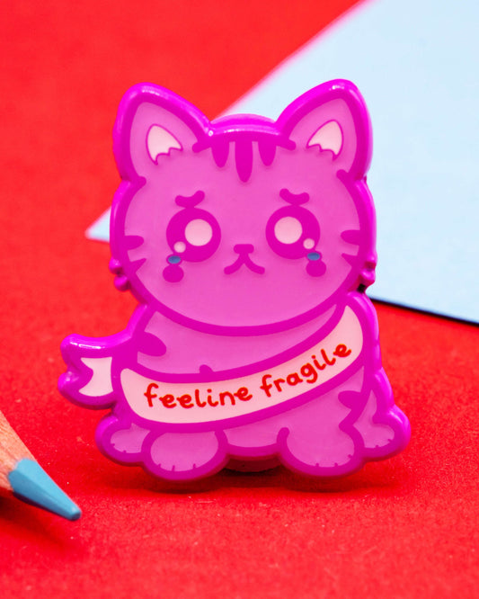 Close-up of the Feeline Fragile enamel pin by Innabox on red background with pencils. The pink cat design looks sad and adorable, wearing a ribbon that says “feeline fragile.” Sold by Rebel Romance, a small UK brand for alternative fashion and thoughtful gifts.