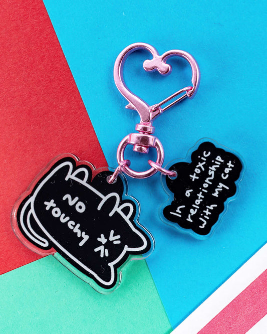 No Touchy Cat Keyring by Innabox, featuring two black acrylic charms with white illustrations. One reads “No Touchy” with a cartoon cat, the other says “In a toxic relationship with my cat.” Attached to a pink heart-shaped metal keyring. Sold by Rebel Romance, a UK indie brand known for quirky accessories and alternative fashion.