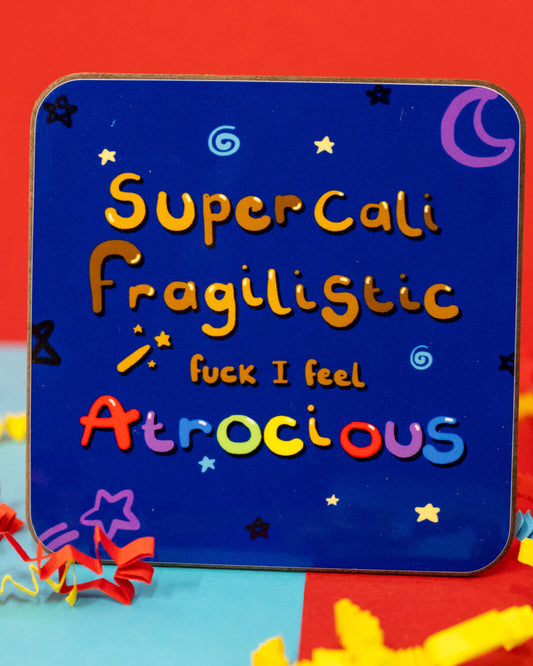 A square wooden coaster with rounded corners featuring the phrase “SuperCaliFragilistic fuck I feel Atrocious” in colourful, playful lettering on a dark blue background with stars, moons, and sparkles. Designed by Innabox, sold by Rebel Romance, a UK small business celebrating alternative and millennial aesthetics.
