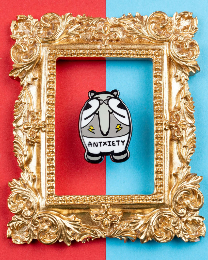 Quirky enamel anteater pin with "ANTXIETY" text displayed inside a gold ornate frame. The anteater has worried eyes and yellow lightning bolts on its belly. Background features a bold half-red, half-blue contrast. Sold by Rebel Romance, a UK brand celebrating alternative style, neurodiversity, and mental health awareness through fun enamel pins and slow fashion.
