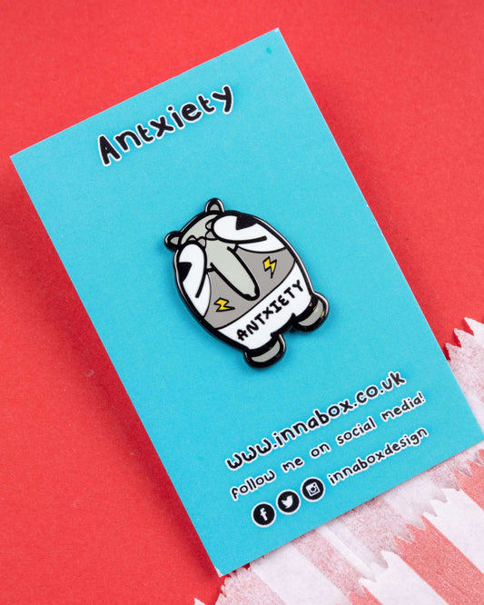 Cute enamel pin on blue backing card featuring a grey anteater with anxious eyes and lightning bolt details on its belly. The word "ANTXIETY" is written across its body. The pin is displayed on red and blue card with striped paper in the corner. Sold by Rebel Romance, a small UK business specialising in alternative fashion, gifts, and mental health-themed accessories.