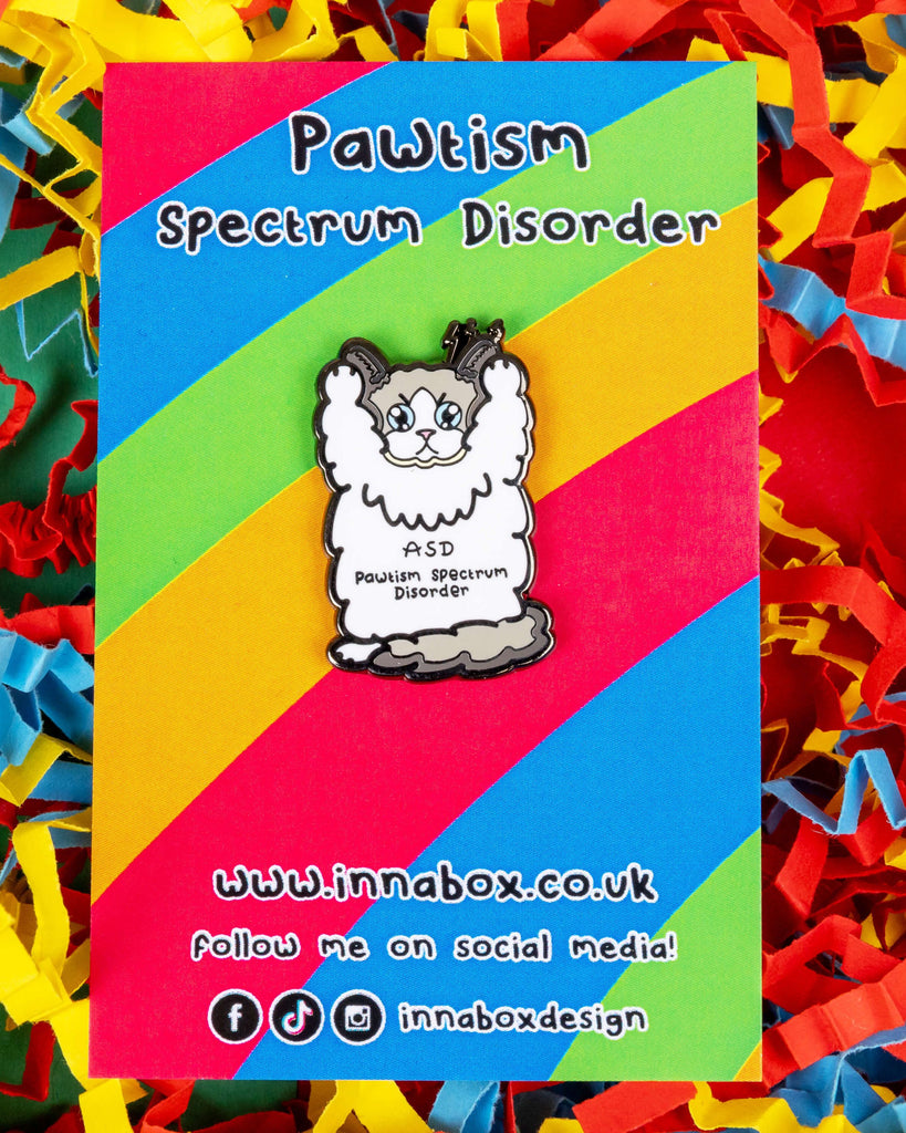 Pawtism Spectrum Disorder cat enamel pin displayed on a colourful rainbow backing card. The design features a fluffy white cat with "ASD Pawtism Spectrum Disorder" written on its body. Designed by Innabox, an alternative UK brand that promotes awareness with fun, inclusive accessories.