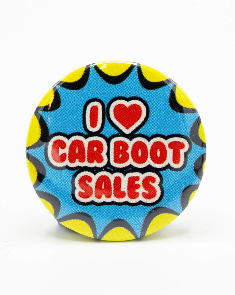 Round button pin badge with a bright comic book-style design that says “I ❤️ Car Boot Sales” in bold red and white lettering on a blue background with yellow and black scalloped edging. Designed by Bite Your Granny, sold by Rebel Romance, a UK small biz celebrating quirky and alternative fashion, gifts, and homeware.