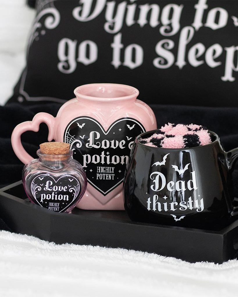 A gothic pink heart‑shaped ceramic mug from Rebel Romance with “Love Potion Highly Potent” printed on the front in a black heart label, styled alongside a black “Dead Thirsty” mug and a small potion bottle. Alternative homeware gift for millennials who love spooky and magical aesthetics.