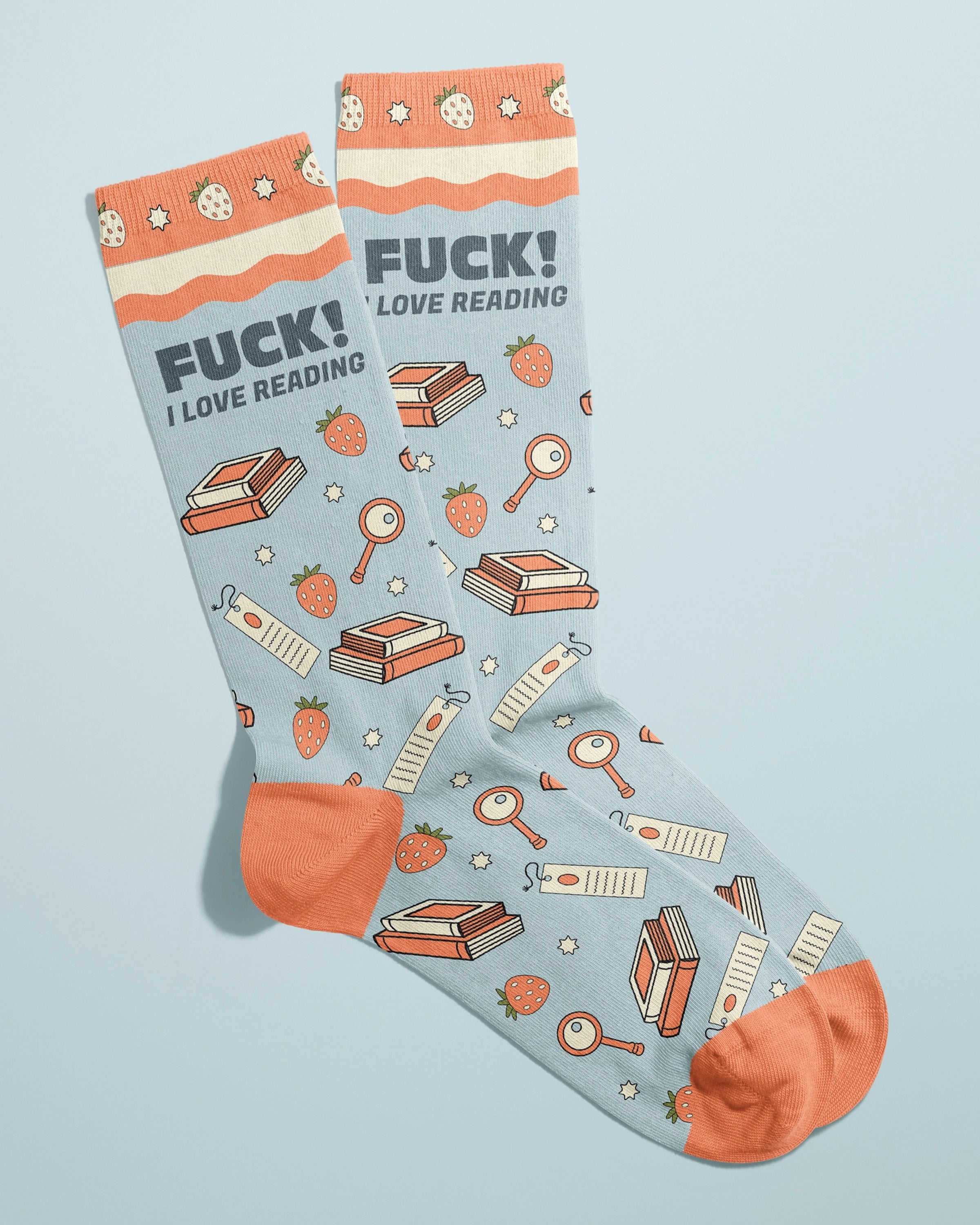 Rebel Romance novelty socks by FUN CLUB laid side-by-side with visible slogan “FUCK! I LOVE READING” in bold lettering. Socks feature strawberries, books, bookmarks, magnifying glasses, and stars on a pastel blue background with orange cuffs and toes.