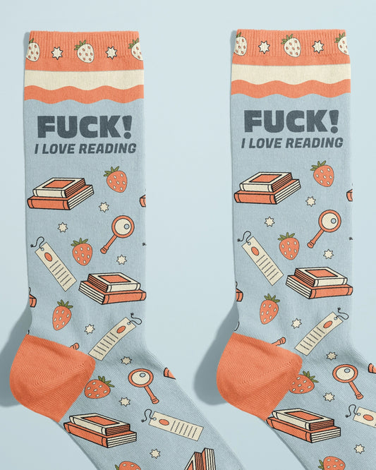 Flat lay of Rebel Romance novelty socks by FUN CLUB with the slogan “FUCK! I LOVE READING,” featuring a pastel blue background, orange toe and cuff, and illustrations of books, strawberries, bookmarks, and stars. Ethically made and perfect for book lovers who enjoy bold, funny fashion.