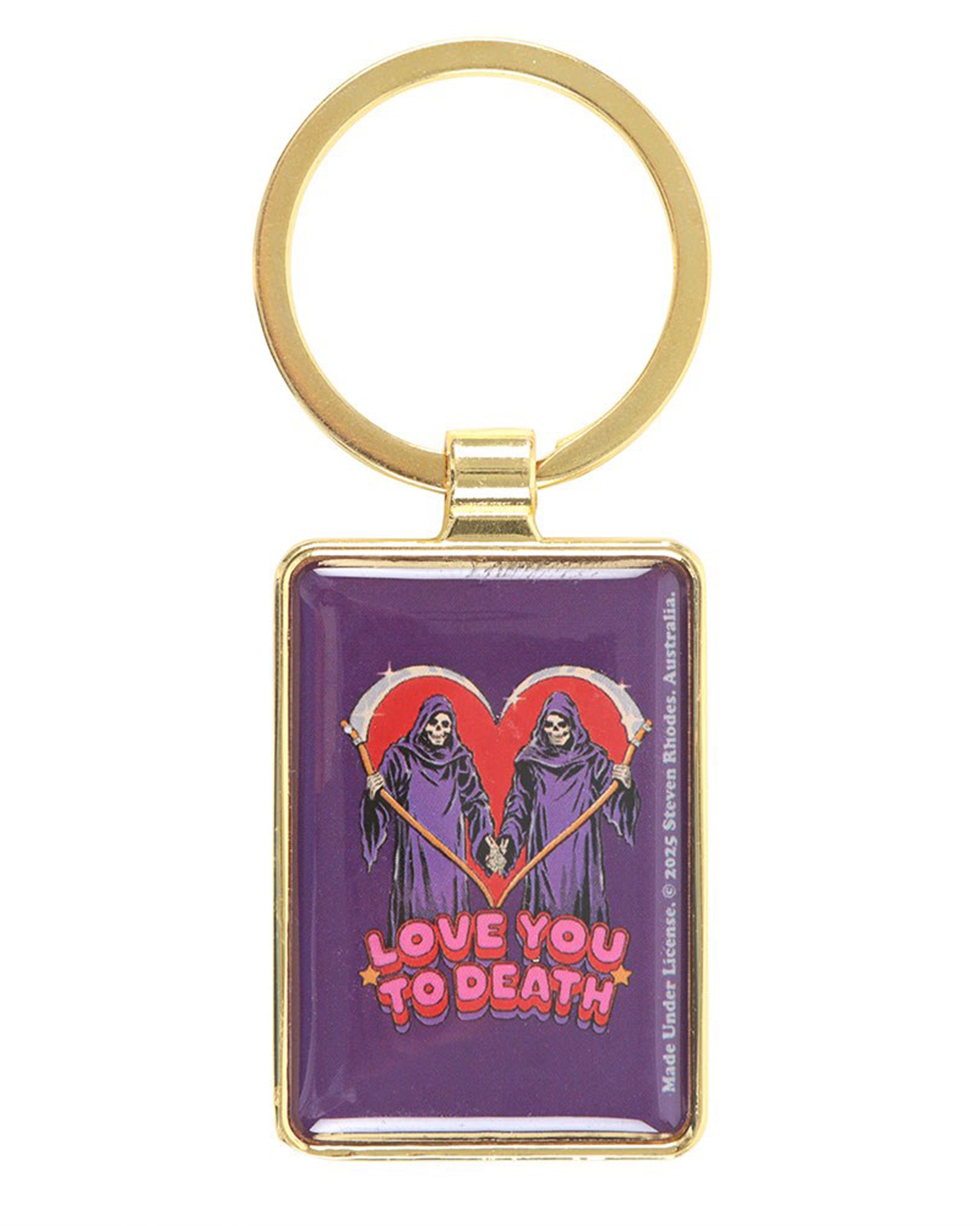 Steven Rhodes “Love You To Death” enamel keyring sold by Rebel Romance. Features two grim reapers in front of a red heart, holding scythes and hands, with text below. Framed in gold metal with a circular keyring attachment. Fun gothic accessory gift.
