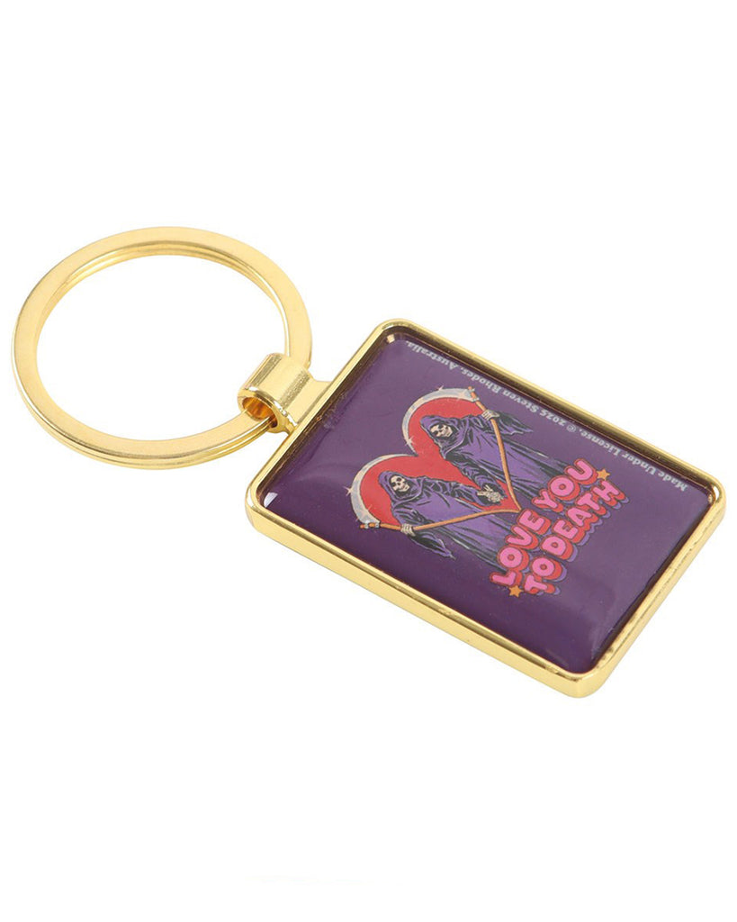 Side angle of the Rebel Romance Steven Rhodes “Love You To Death” keyring showing its gold metal frame and enamel design. Features two grim reapers with scythes forming a red heart, above retro lettering. Perfect gothic keychain for alternative fashion fans.