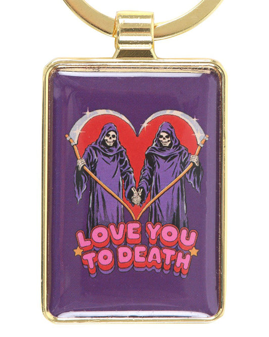 Rectangular enamel keyring by Rebel Romance featuring Steven Rhodes’ “Love You To Death” design. Two grim reapers in purple robes hold scythes forming a red heart shape with bold retro text. Gold metal frame and keyring loop. Alternative gothic accessory for dark humour lovers.