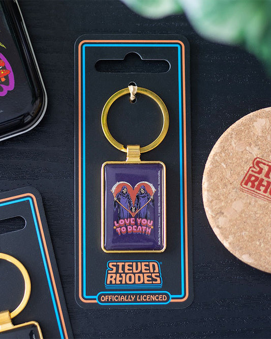 Lifestyle shot of the Rebel Romance Steven Rhodes “Love You To Death” enamel keyring in packaging, placed on a black surface beside other accessories. Features grim reapers with scythes in a heart shape. Officially licensed alternative gothic keychain.