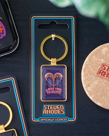 Lifestyle shot of the Rebel Romance Steven Rhodes “Love You To Death” enamel keyring in packaging, placed on a black surface beside other accessories. Features grim reapers with scythes in a heart shape. Officially licensed alternative gothic keychain.