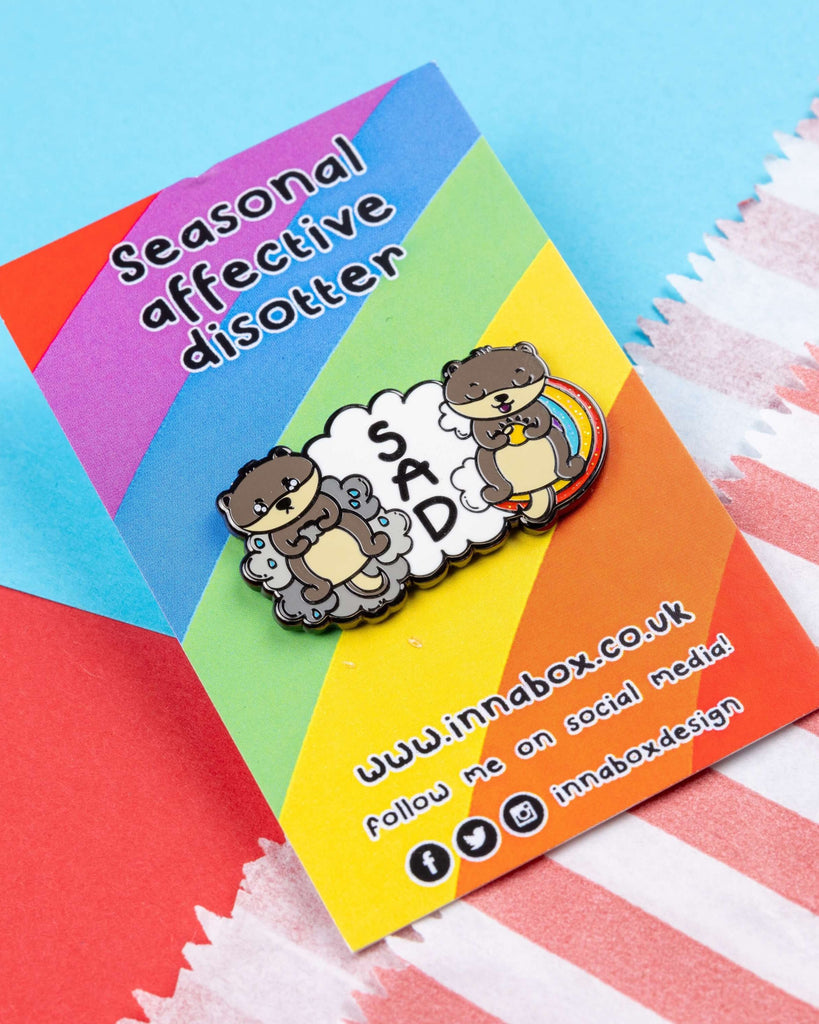 Seasonal Affective Disotter enamel pin by Rebel Romance, featuring two cartoon otters—one sad on a grey cloud, one happy on a rainbow—with the word “SAD” between them, designed by Innabox. The pin is displayed on colourful rainbow backing card with bold text. Ideal for mental health awareness and cute pin collectors.
