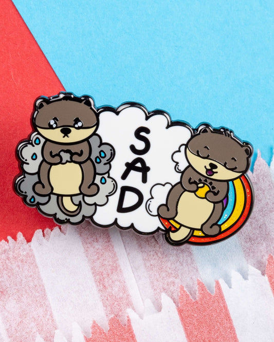 Close-up of the Seasonal Affective Disotter glittery enamel pin by Rebel Romance. The hard enamel pin shows a sad otter on a grey cloud and a cheerful otter on a rainbow with the letters “SAD” in the middle. A playful design to support awareness of seasonal affective disorder, created by Innabox.