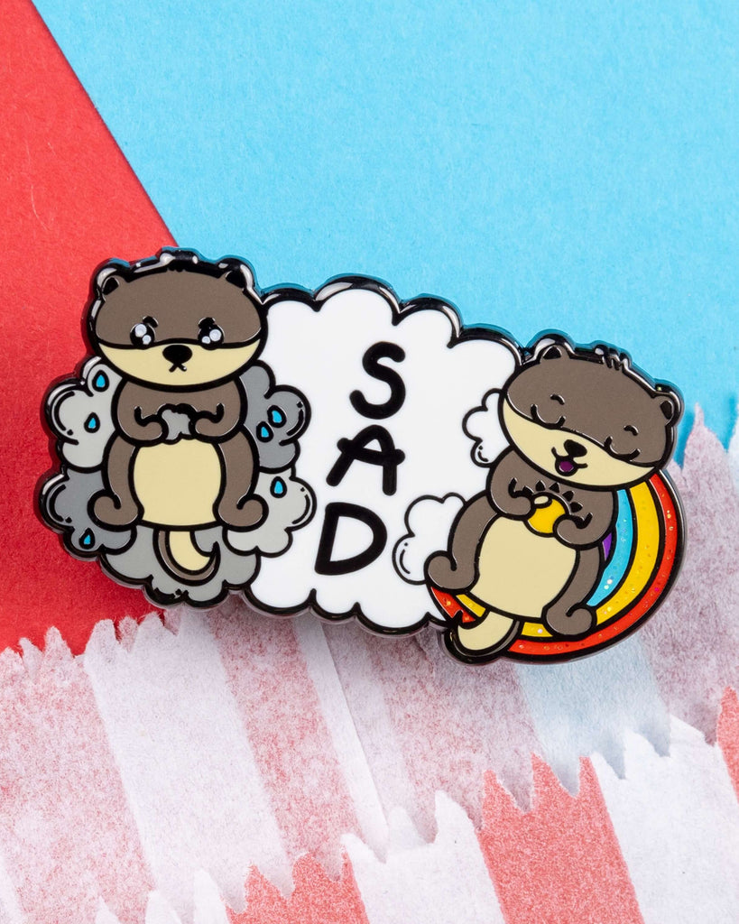 Close-up of the Seasonal Affective Disotter glittery enamel pin by Rebel Romance. The hard enamel pin shows a sad otter on a grey cloud and a cheerful otter on a rainbow with the letters “SAD” in the middle. A playful design to support awareness of seasonal affective disorder, created by Innabox.
