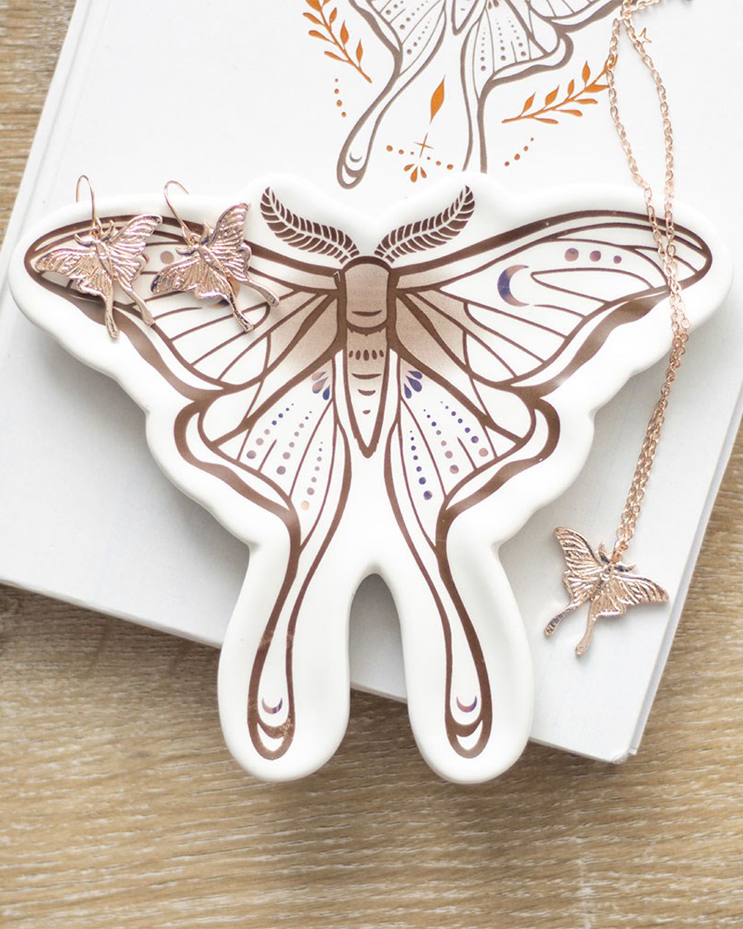 The Rebel Romance luna moth ceramic trinket dish styled with matching moth jewellery on a book, creating an ethereal display for storing earrings, necklaces or rings.