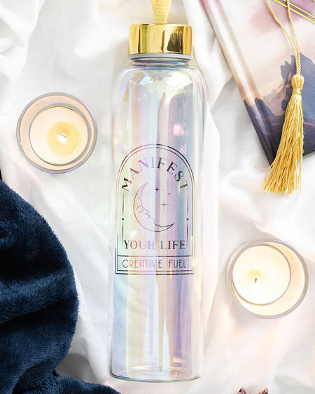 Flat lay of the Manifest Your Life iridescent water bottle surrounded by cosy elements: candles, a navy blanket, and a tasselled journal. The bottle’s pastel glow and gold lettering create a calming, mystical vibe.