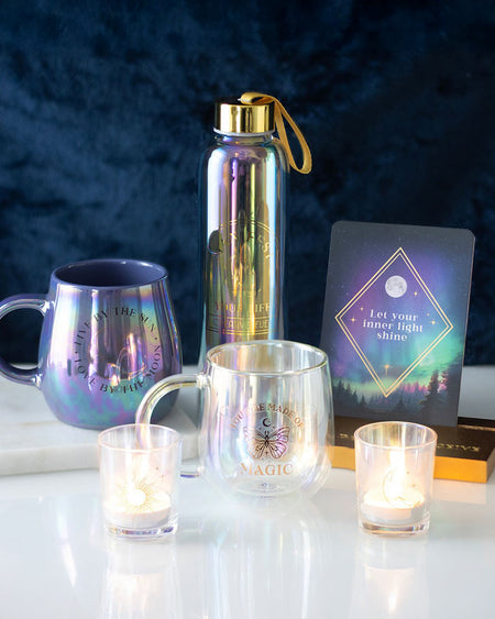 A dreamy, iridescent glass water bottle from Rebel Romance with "Manifest Your Life – Creative Fuel" in elegant gold lettering. Styled with mystical mugs, candles, and an inspirational card against a deep blue velvet background. Perfect for adding a magical touch to your hydration routine.
