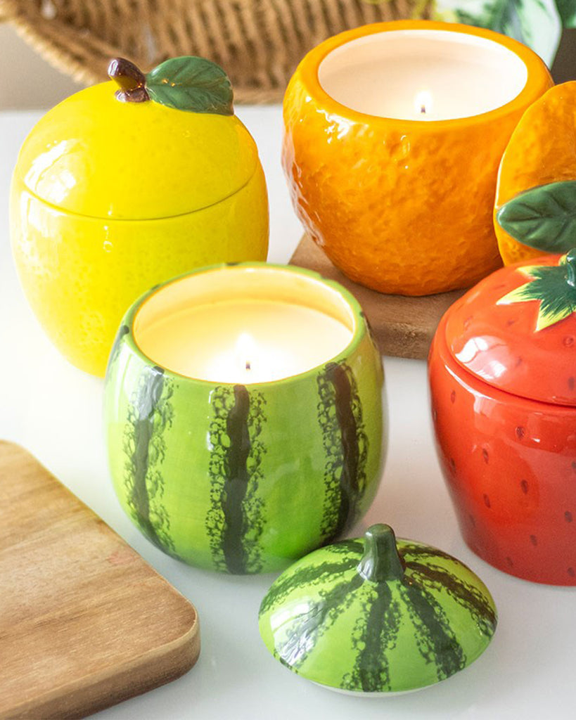 A collection of fruit-shaped candle jars, featuring watermelon, lemon, orange, and strawberry designs. The green watermelon jar with a realistic rind pattern and removable lid is in focus, with the candle lit inside.