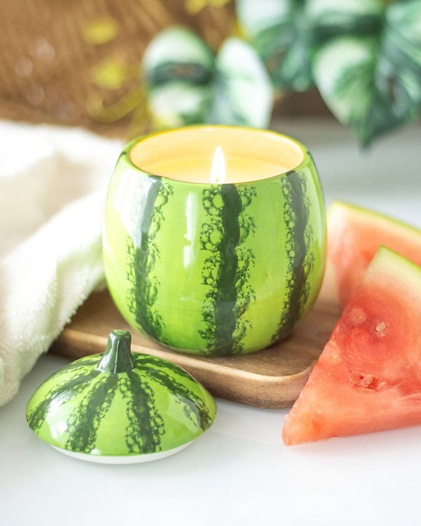 The watermelon candle jar lit, casting a warm and inviting glow. The ceramic lid with a green stem rests beside it, showing off the realistic summer fruit design.