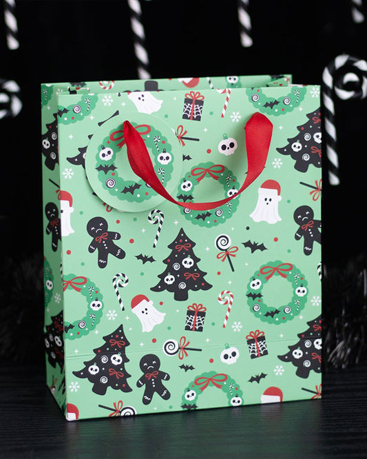 Lifestyle image of the Rebel Romance Merry Creepmas gift bag placed on a black wooden surface. The mint green bag is covered in spooky Christmas illustrations like ghosts, creepy trees, skulls, and festive sweets, perfect for alternative holiday gifting.
