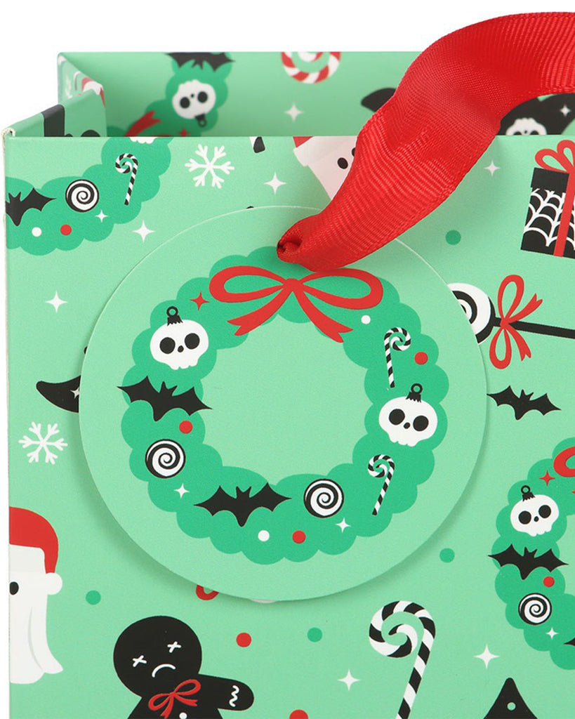 Close-up of the Merry Creepmas gift bag by Rebel Romance showing the round green wreath gift tag decorated with skulls, bats, candy canes and a red bow, attached with red ribbon handles. The background pattern includes spooky Christmas-themed illustrations.