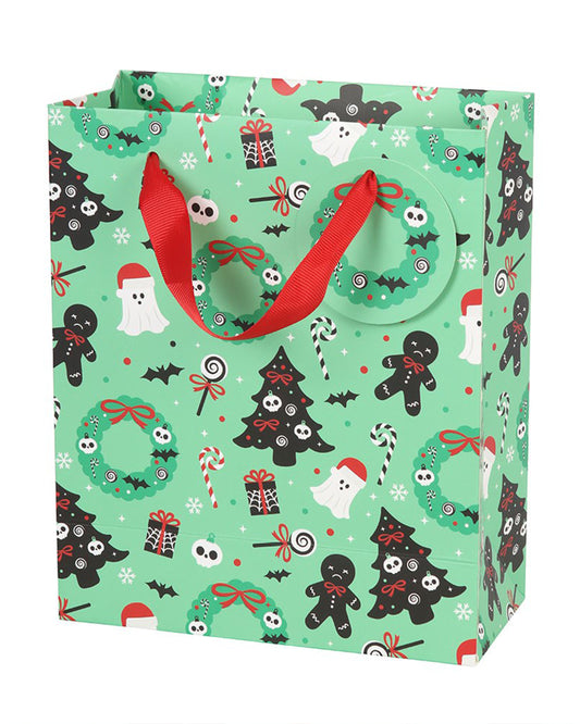 Mint green Merry Creepmas gift bag by Rebel Romance featuring spooky festive illustrations including black Christmas trees with skull baubles, creepy gingerbread men, candy canes, ghosts in Santa hats, bats, wreaths, skulls, and presents. Includes red ribbon handles and a matching wreath-shaped gift tag.