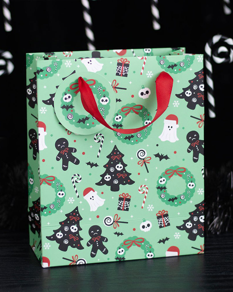 Lifestyle image of the Rebel Romance Merry Creepmas gift bag placed on a black wooden surface. The mint green bag is covered in spooky Christmas illustrations like ghosts, creepy trees, skulls, and festive sweets, perfect for alternative holiday gifting.