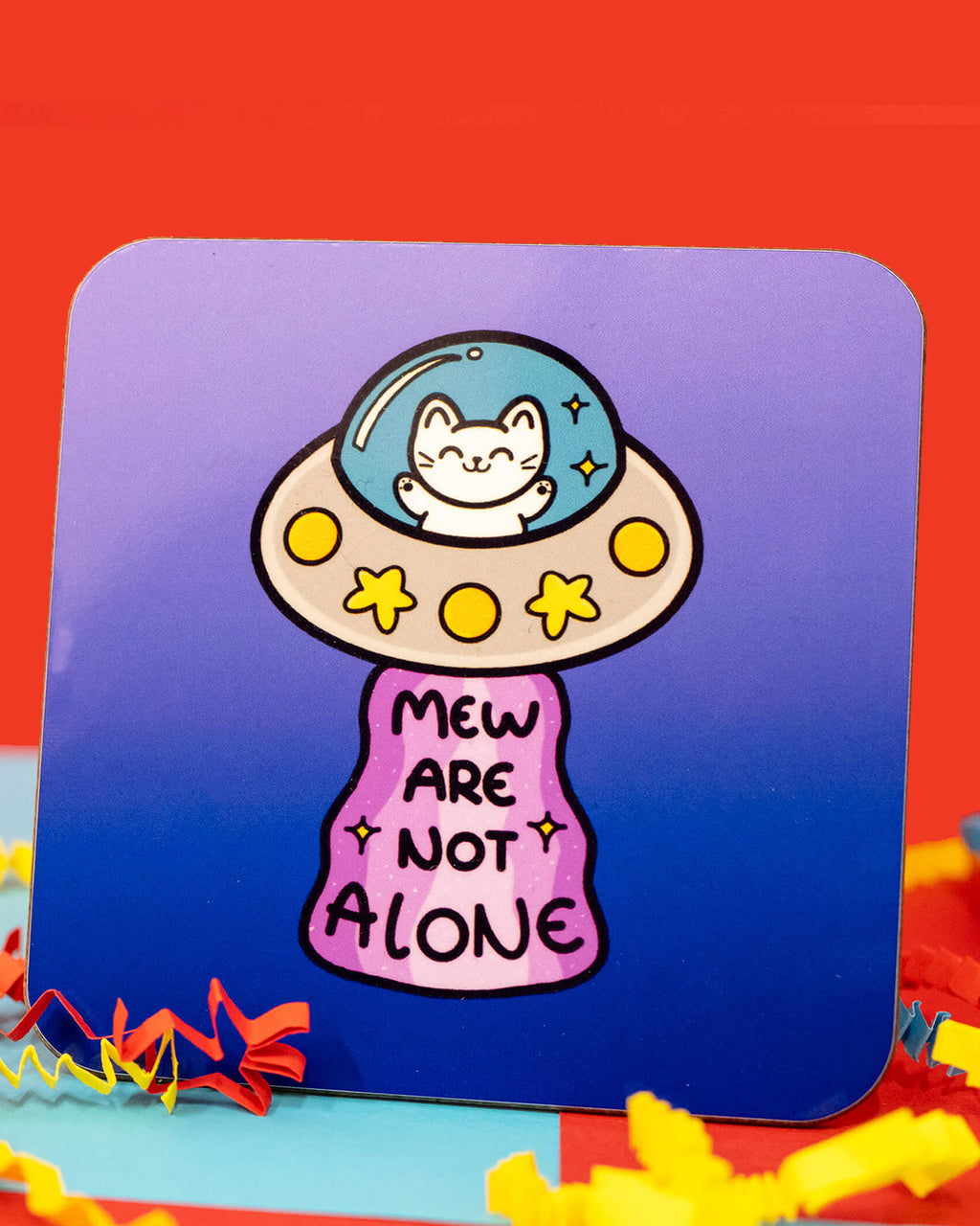 Close-up of the Cat UFO Mew Are Not Alone Coaster by Innabox. The square coaster has rounded corners and a gradient blue background. The design features a cute white cat sitting inside a grey UFO with yellow lights and star details. A pink sparkly beam shines down with the words 