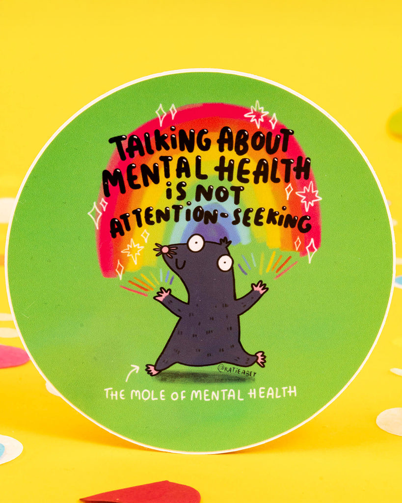 A round vinyl sticker by Katie Abey, available from Rebel Romance. Features a cartoon mole with wide eyes standing under a rainbow with the text “Talking about mental health is not attention-seeking.” A bold and supportive message with a playful design, ideal for laptops, notebooks, or gifts promoting mental health awareness.