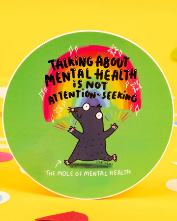 A round vinyl sticker by Katie Abey, available from Rebel Romance. Features a cartoon mole with wide eyes standing under a rainbow with the text “Talking about mental health is not attention-seeking.” A bold and supportive message with a playful design, ideal for laptops, notebooks, or gifts promoting mental health awareness.