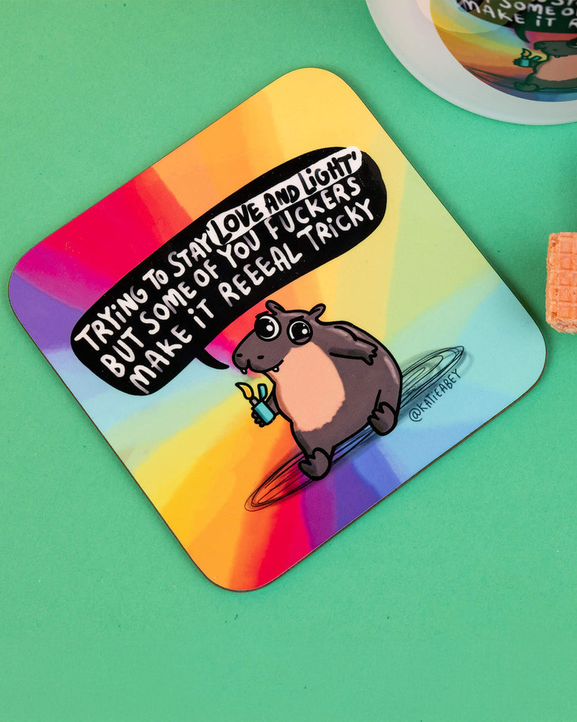 Flat lay of the Moo Deng Hippo Coaster featuring the same rainbow design and sassy hippo illustration, placed next to a matching mug and pink wafer biscuits on a mint green and yellow background. Bold, funny giftware by Katie Abey.