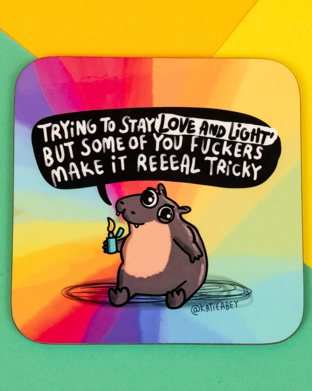 Front view of the Moo Deng Hippo Coaster on a flat surface. The coaster shows a moody hippo holding a lighter with a speech bubble that reads “Trying to stay love and light but some of you f*ckers make it reeeal tricky”, set against a bold rainbow backdrop. Designed by Katie Abey.