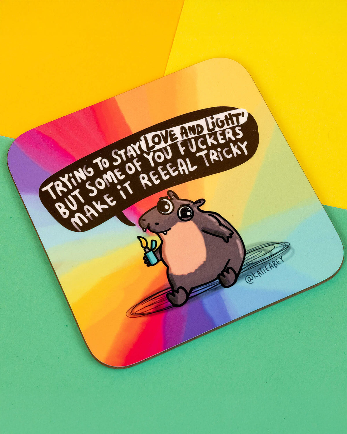 Close-up angled shot of the Moo Deng Hippo Coaster designed by Katie Abey, featuring a grumpy grey hippo holding a lighter and saying “Trying to stay love and light but some of you f*ckers make it reeeal tricky”, on a vibrant rainbow background. Shown on a colourful green and yellow surface.